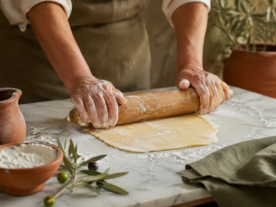 Flour-dusted hands rolling fresh pasta dough with a wooden pin on a marble counter