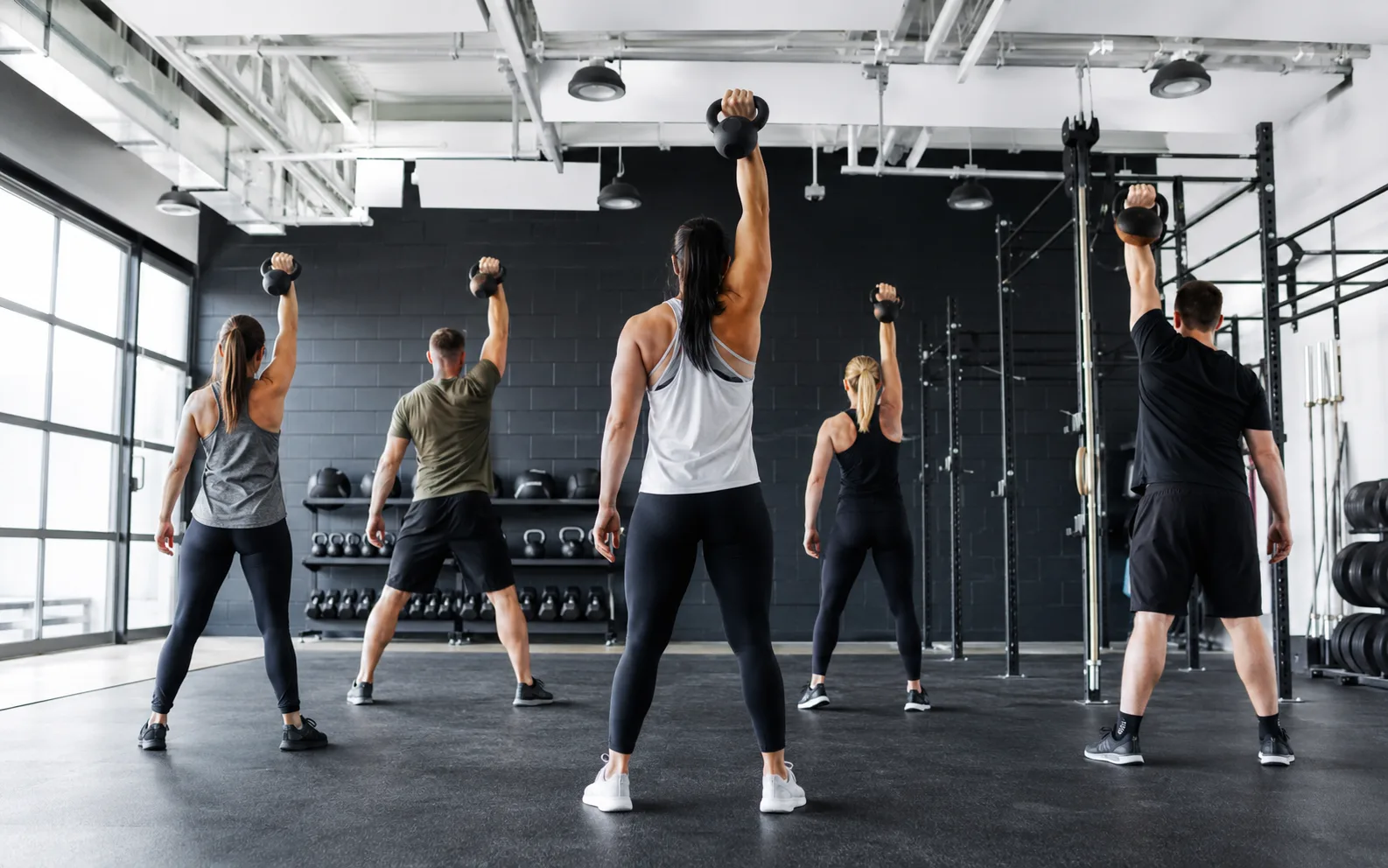 Five members mid-class pressing kettlebells overhead in unison, facing a dark brick wall lined with kettlebell racks