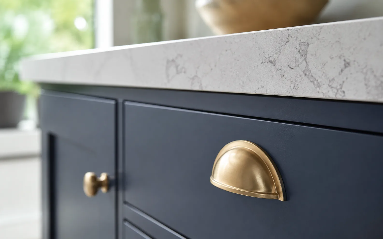 Close-up of a brushed brass cup pull and knob on navy shaker cabinetry, with a quartz countertop edge above